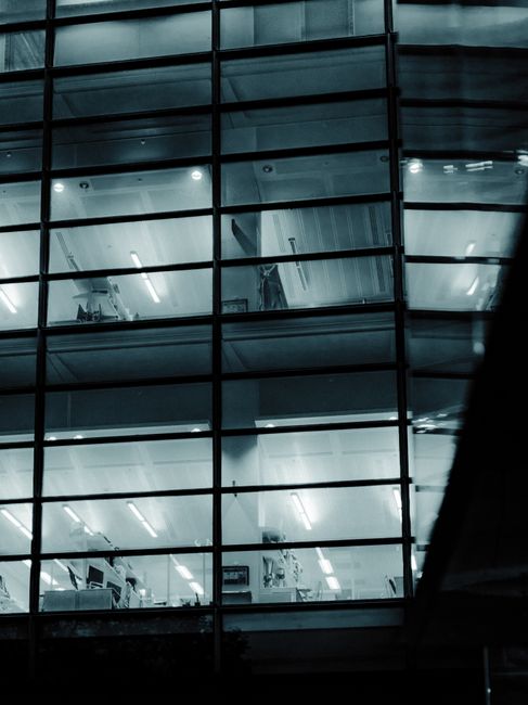 The image shows a modern office building with a reflective glass exterior, situated along a waterway. The building features multiple rectangular glass facades with visible internal structures and a pr