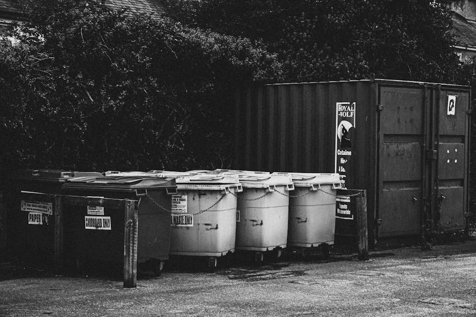 A row of four large white wheelie bins with closed lids, labeled for waste disposal, positioned on a paved area next to a wooden post with a small sign. To the right of the wheelie bins is a large met