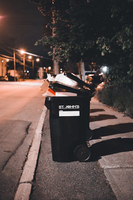 A row of four large white wheelie bins with closed lids, labeled for waste disposal, positioned on a paved area next to a wooden post with a small sign. To the right of the wheelie bins is a large met